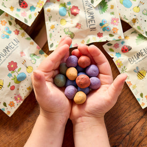 Les mains des enfants tiennent des bombes à graines colorées ; à l'arrière-plan, des sacs à motifs floraux reposent sur une table en bois.