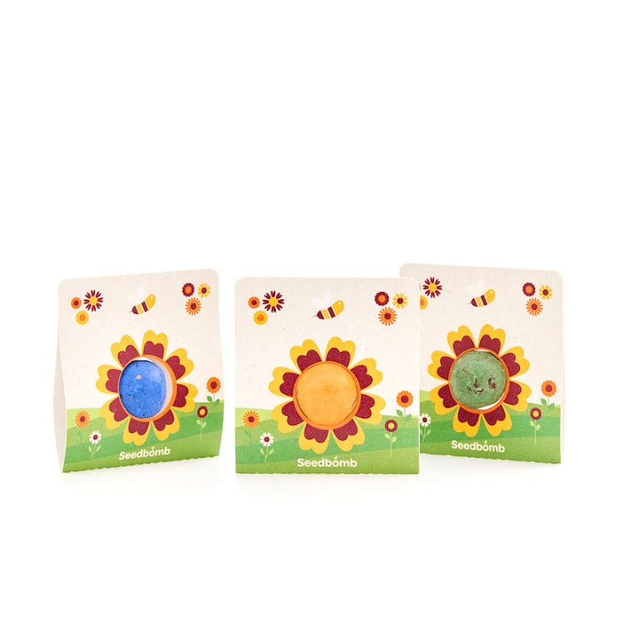 Trois bombes de graines colorées dans un emballage avec des motifs de tournesol et d'abeille sur fond blanc.
