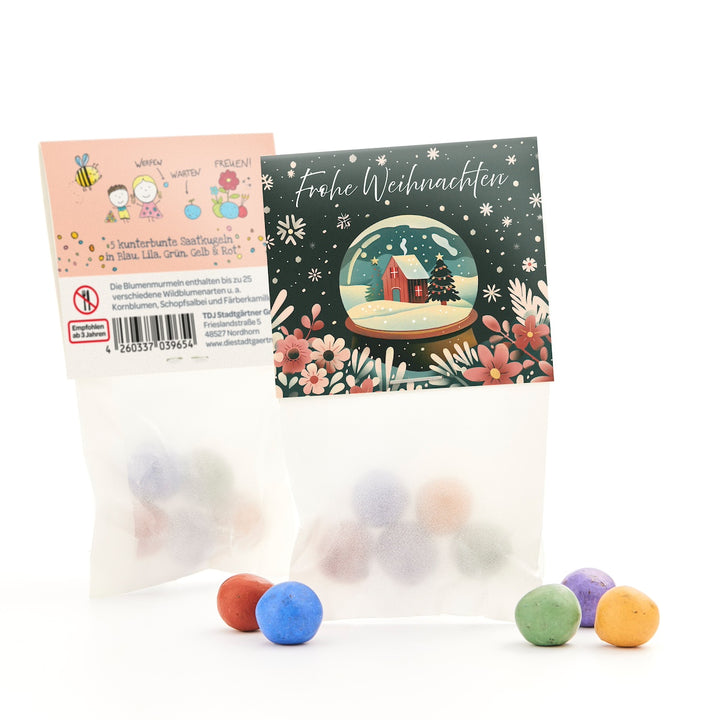 Deux petits sacs de boules de graines colorées, l'un avec une carte de vœux de Noël comportant un motif de boule à neige et l'inscription « Joyeux Noël », à côté des boules colorées sur fond blanc.