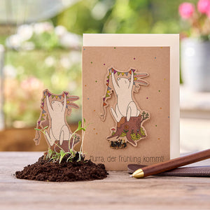 Carte de vœux avec illustration d'un lapin joyeux tenant un fanion sur une souche d'arbre ; texte : « Hourra, le printemps arrive ! » ; petites plantes dans le sol au premier plan.