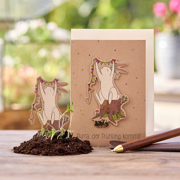 Carte de vœux avec illustration d'un lapin joyeux tenant un fanion sur une souche d'arbre ; texte : « Hourra, le printemps arrive ! » ; petites plantes dans le sol au premier plan.