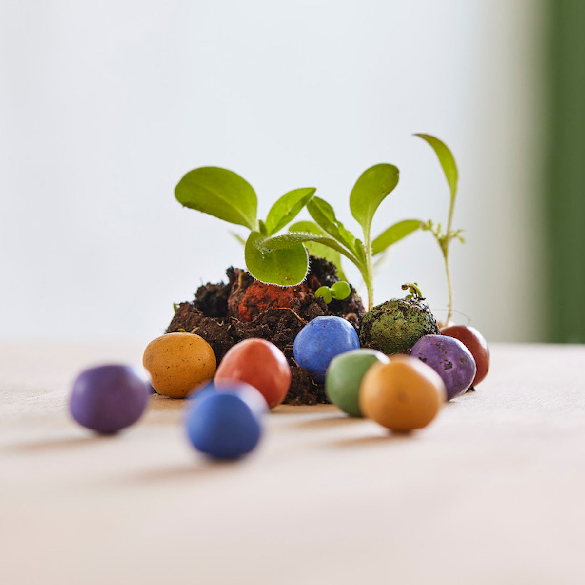 Plusieurs bombes de graines colorées et petites plantes vertes sur un fond clair.