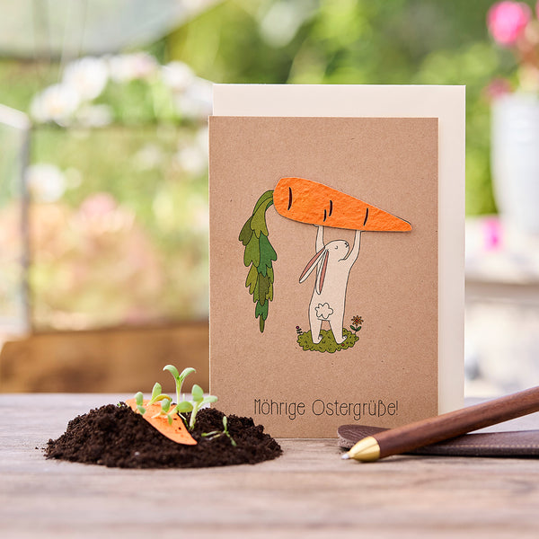 Carte de Pâques avec un lapin dessiné tenant une grosse carotte, à côté de lui de la terre avec de petites plantes et une décoration de carotte, texte "Carottey Easter greetings!".