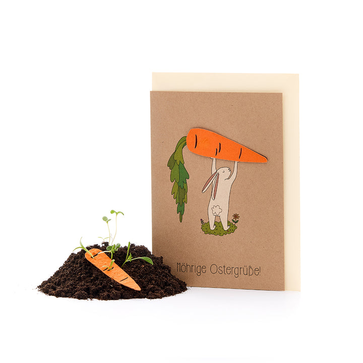 Une carte de Pâques avec le dessin d'un lapin tenant une grosse carotte au-dessus de sa tête, à côté d'un petit monticule de terre contenant une carotte en papier et des pousses de plantes. Le texte dit : « Vœux de Pâques carotte ! »
