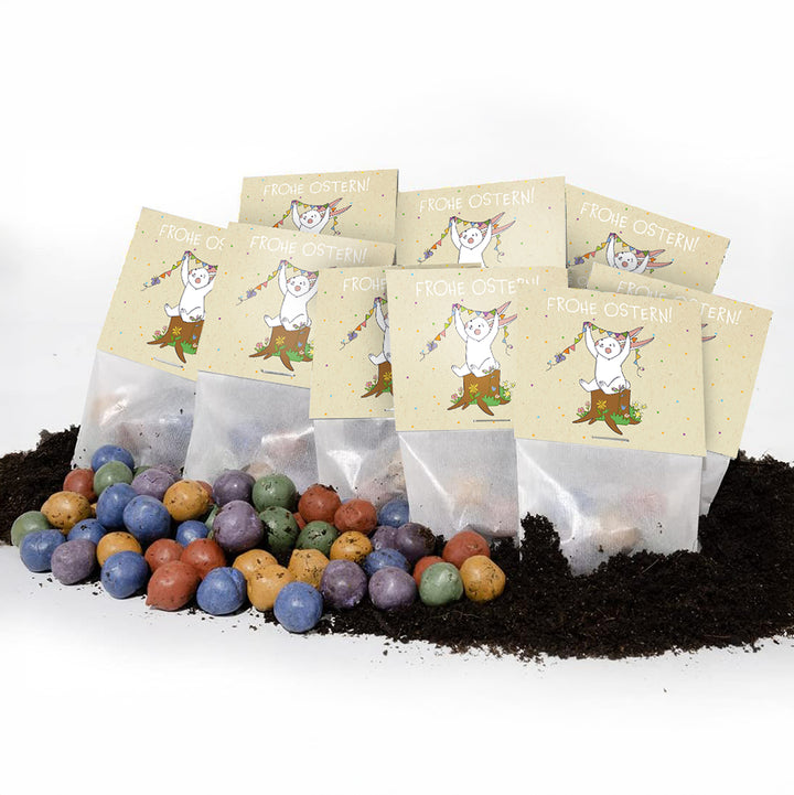 Des bombes de graines colorées reposent sur le sol, derrière elles plusieurs petits sacs avec les mots « Joyeuses Pâques ! » et un motif de lapin.