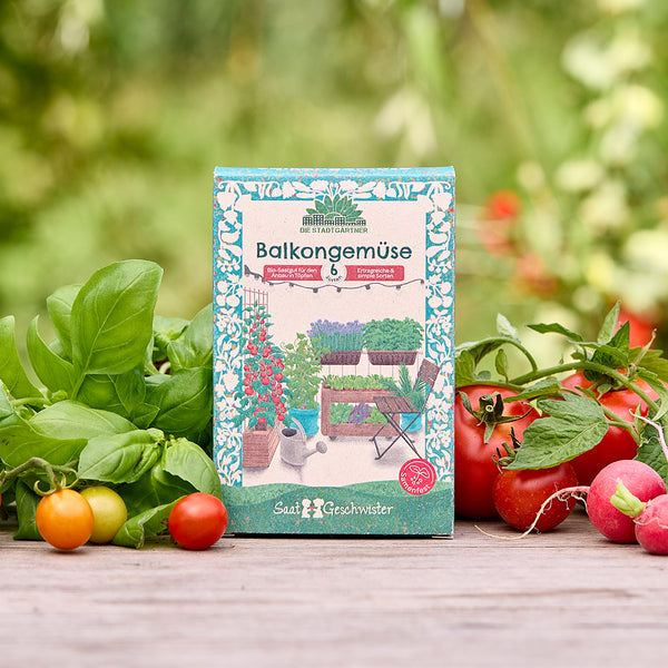 Paquet de légumes de balcon avec des graines pour divers légumes sur une table en bois, entouré d'herbes fraîches, de tomates et de radis à l'extérieur.