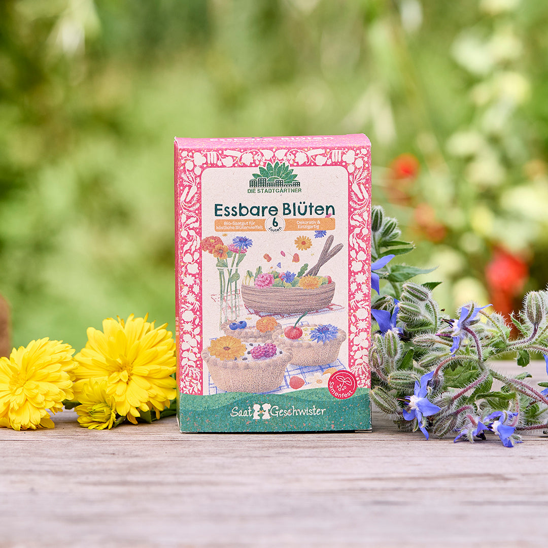 Un sachet de graines étiqueté « Fleurs comestibles » est posé sur une table en bois, entouré de fleurs jaunes et violettes, sur un fond vert flou.