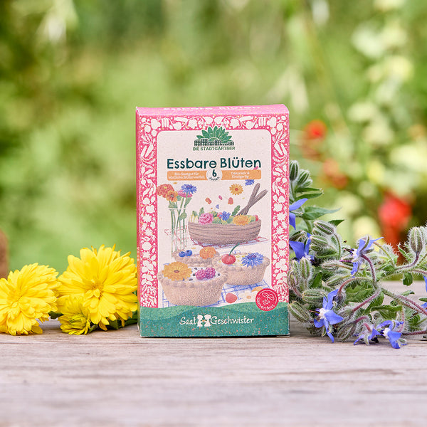 Un sachet de graines étiqueté « Fleurs comestibles » est posé sur une table en bois, entouré de fleurs jaunes et violettes, sur un fond vert flou.