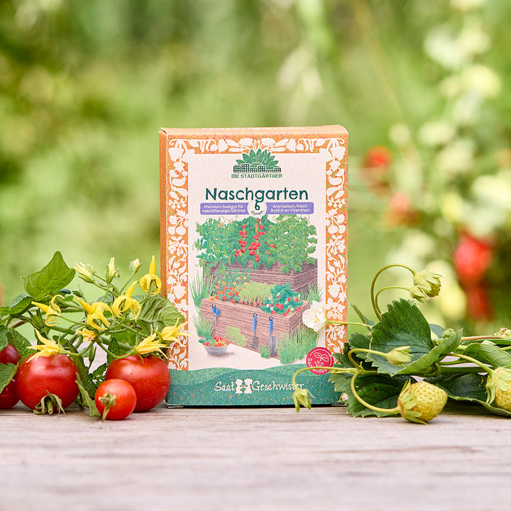 Un paquet de graines « Naschgarten » se trouve sur une table en bois dans le jardin, entouré de tomates fraîches, de fraises et de feuillage vert.