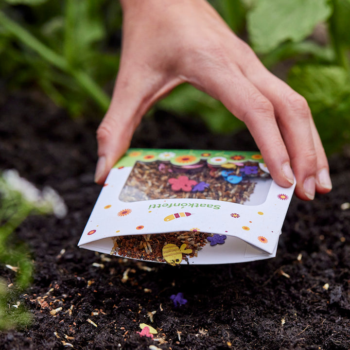 Une main disperse des graines de fleurs provenant d'un sac décoré de couleurs vives sur le sol du jardin.