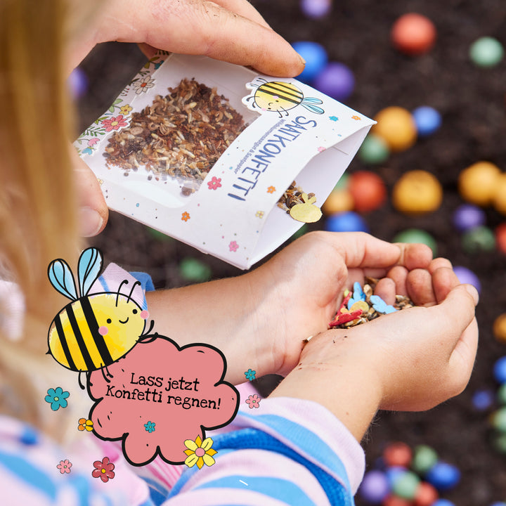Un enfant tient des confettis de graines dans ses mains tandis qu'on verse dessus un sachet de graines florales. Au premier plan, un dessin d'abeille avec la bulle « Qu'il pleuve des confettis ! » est affiché. À l'arrière-plan, des boules colorées sur la terre.