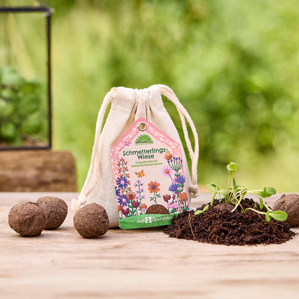 Sac en tissu étiqueté « Prairie aux papillons », à côté des boules de graines et de la terre avec de jeunes plantes, avec une nature floue en arrière-plan.