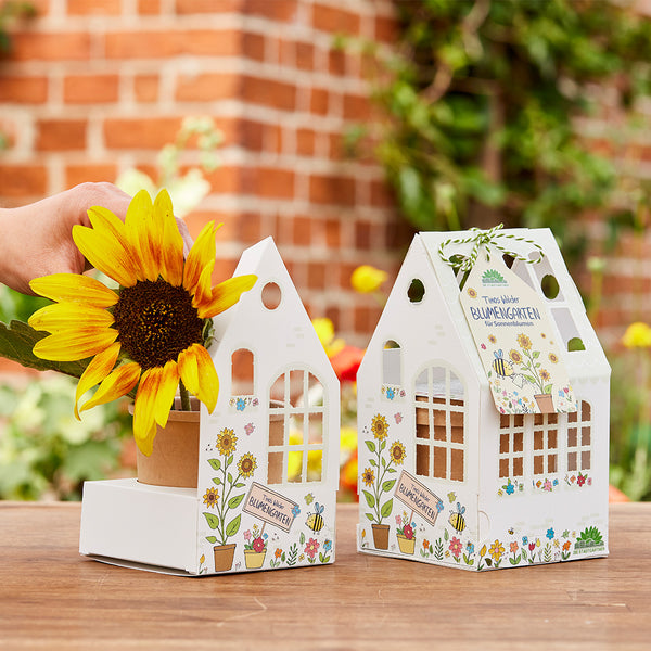 Main avec tournesol et deux paquets décoratifs en forme de maison avec l'inscription "Jardin de fleurs" sur une table en bois à l'extérieur.