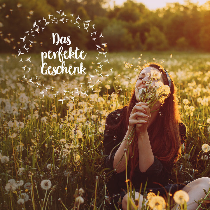 Jeune femme assise dans un pré plein de pissenlits, tenant un bouquet de pissenlits et souriant, entourée de graines volantes ; texte dans l'image : « Le cadeau parfait. »