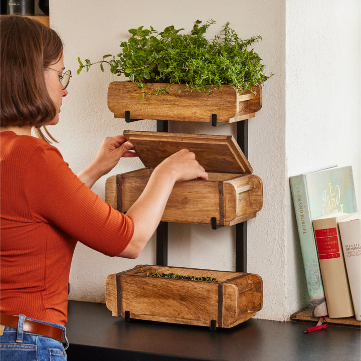 Une femme place une boîte en bois sur une étagère avec des herbes dans une cuisine.