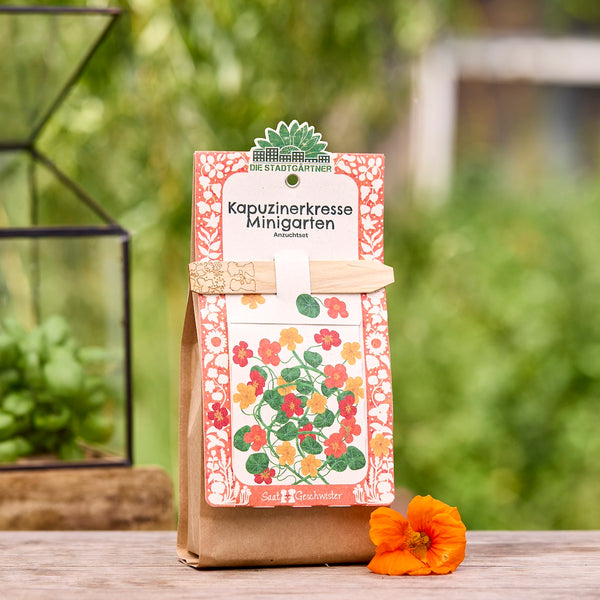 Sac brun contenant un kit de culture de capucine, décoré d'une illustration colorée des fleurs et d'un tuteur en bois, dans le jardin sur une table en bois, à côté d'une fleur de capucine orange.