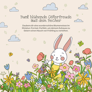 Illustration d'un lapin blanc dans une prairie colorée avec des fleurs printanières, des papillons et des nuages ; en haut se trouve un texte allemand sur la joie de Pâques et la décoration florale.