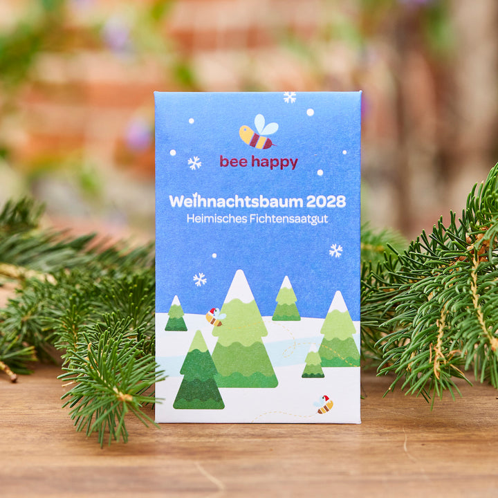 Paquet avec l'inscription « Christmas Tree 2028 Native Spruce Seed » et un motif coloré d'abeille et de sapin, décoré de branches de sapin au premier plan.