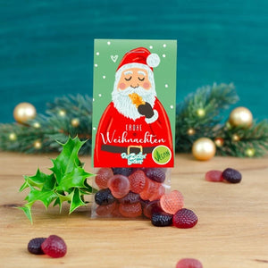 Carte de vœux de Noël avec une image du Père Noël entouré de feuilles de houx et de gommes aux fruits sur une table en bois sur un fond de branches de sapin floues.