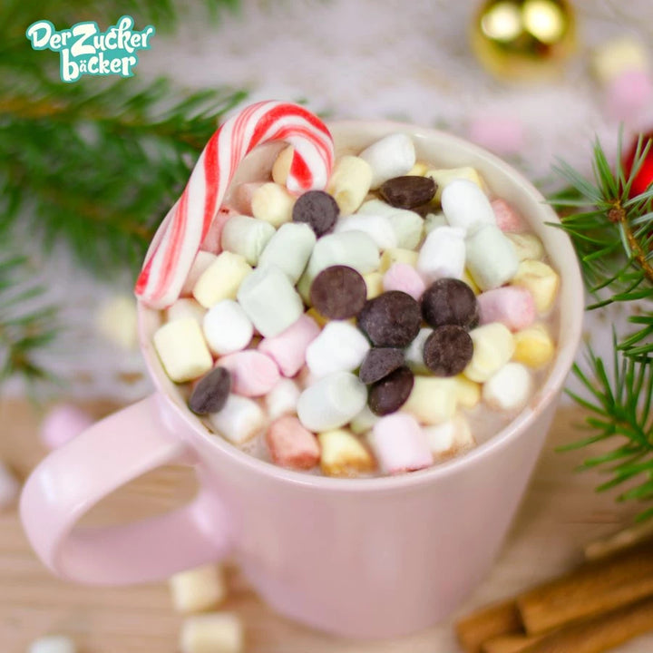 Une tasse de chocolat chaud avec des mini guimauves et des pépites de chocolat, à côté d'une canne en bonbon, entourée de branches de sapin et de décorations de Noël.