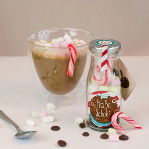 Un verre de chocolat chaud avec des guimauves, à côté une bouteille étiquetée « Chocolat chaud » et des cannes de bonbon, avec des guimauves et des pépites de chocolat éparpillées au premier plan.