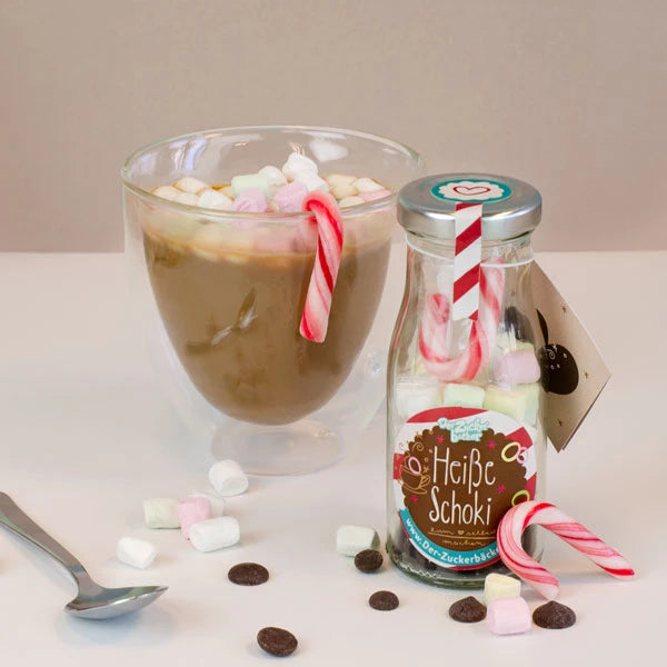 Un verre de chocolat chaud avec des guimauves, à côté une bouteille étiquetée « Chocolat chaud » et des cannes de bonbon, avec des guimauves et des pépites de chocolat éparpillées au premier plan.