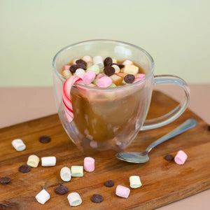 Chocolat chaud dans une tasse transparente avec des guimauves, des pépites de chocolat et de la canne à sucre, servi sur une planche en bois.