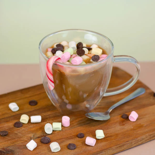 Chocolat chaud dans une tasse transparente avec des guimauves, des pépites de chocolat et de la canne à sucre, servi sur une planche en bois.