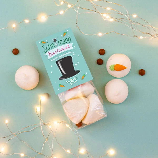 Emballage avec des guimauves "kit bonhomme de neige", entourées de petites boules de chocolat et de guirlandes lumineuses sur fond vert menthe.