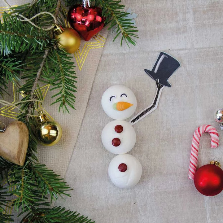 Un bonhomme de neige fait de guimauves avec un pain de sucre, des gouttes de chocolat et un nez de carotte repose sur un tissu à côté de décorations de Noël et de branches de sapin.