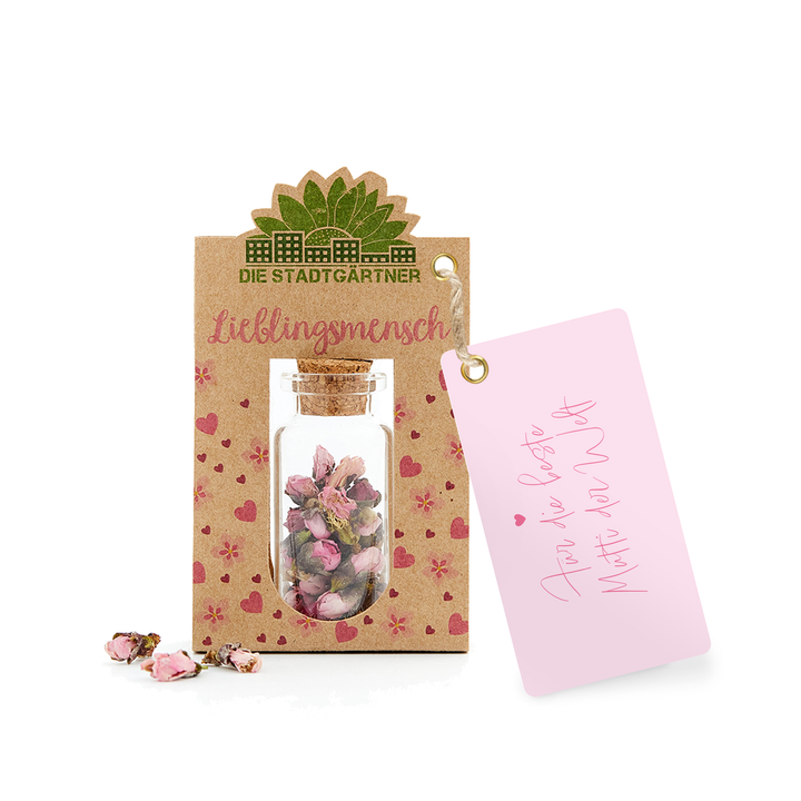 Étiquette cadeau avec l'inscription « Personne préférée » et une petite bouteille en verre avec des fleurs séchées sur un fond de papier kraft à côté d'une carte rose avec un cœur et « Pour toi ».