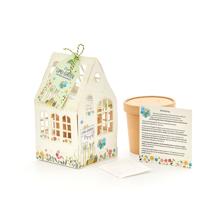 Un kit de bricolage pour que les enfants puissent planter de la mélisse, composé d'une maison décorative en carton, d'un pot de fleurs, d'un sachet de graines et d'instructions.