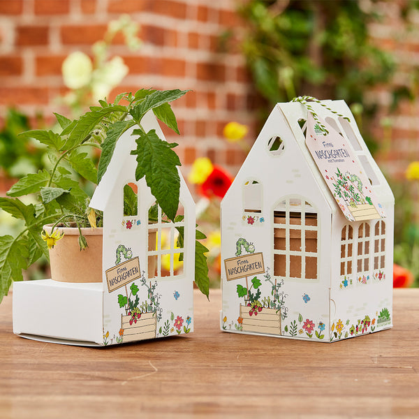 Deux petits paquets en forme de maison étiquetés « Finn's Sweet Garden », l'un contenant une plante en pot, avec des fleurs floues et un mur de briques en arrière-plan.