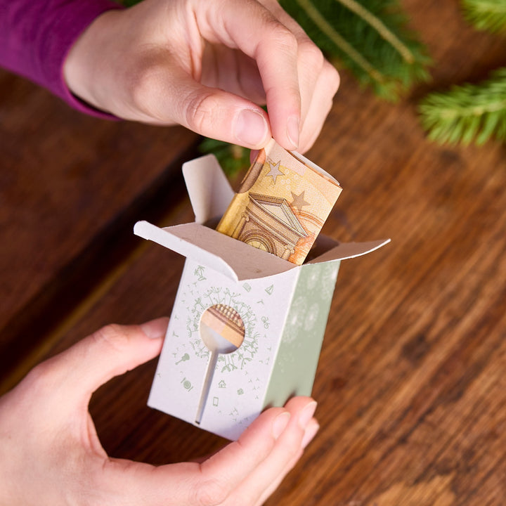 Des mains placent un billet d'euro plié dans une petite boîte cadeau en papier sur une table en bois.