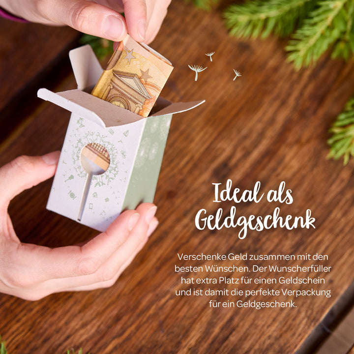 Des mains placent un billet de banque plié dans une petite boîte cadeau en papier, avec le texte « Idéal comme cadeau en espèces » écrit à côté, sur une table en bois avec des branches de sapin.