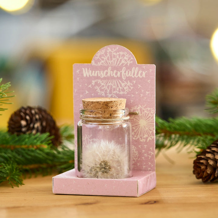 Un petit pot en verre avec un bouchon en liège, contenant un pissenlit, se dresse sur un emballage rose avec l'inscription « Wish Fulfiller », décoré de branches de sapin et de cônes.