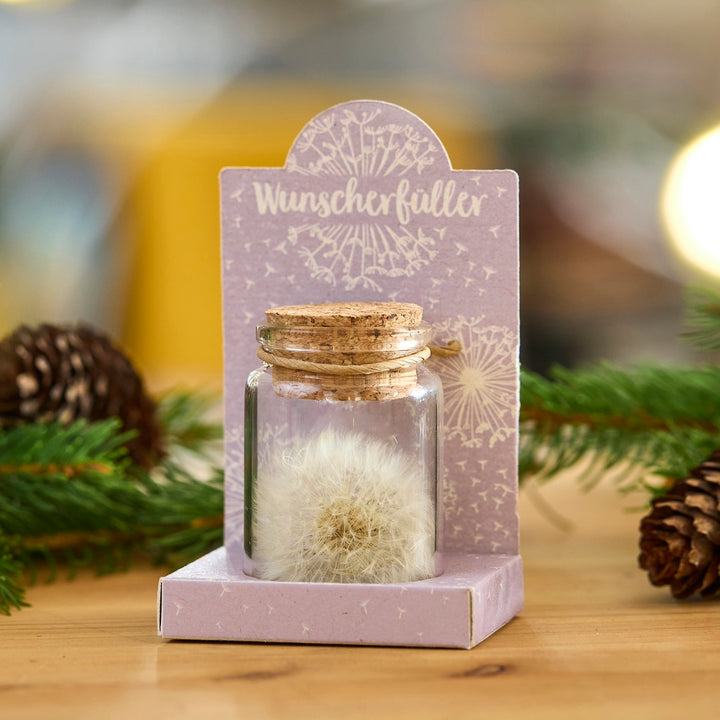 Bouteille en verre avec un pissenlit et un bouchon en liège, sur un emballage violet avec l'inscription "Wish Fulfiller", décoré de branches de sapin et de cônes.