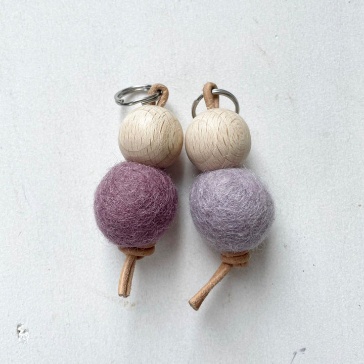 Deux porte-clés faits main avec des boules en bois et des boules en feutre de laine violettes sur fond blanc.