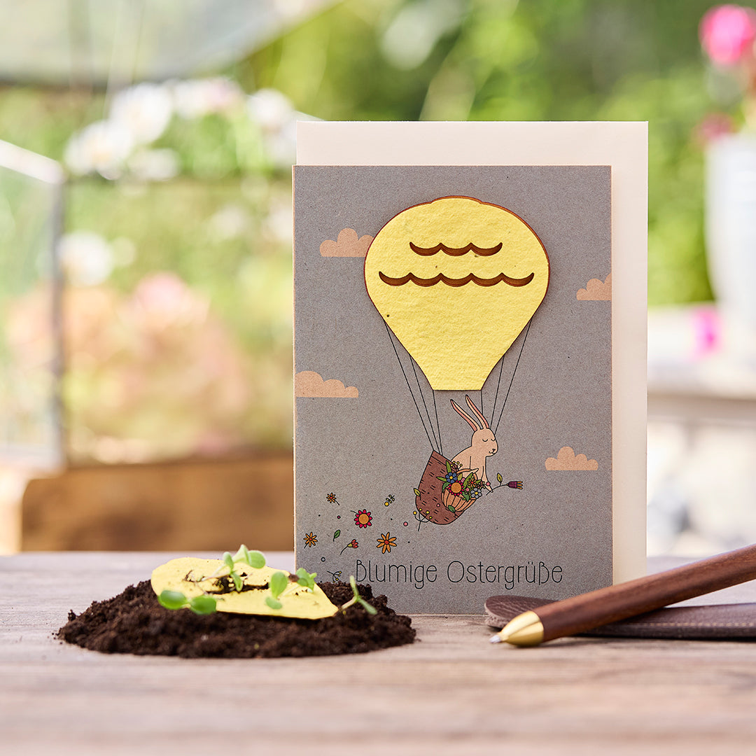 Carte de Pâques avec un lapin dans une montgolfière, à côté de lui de la terre avec des plantes en germination et un stylo.