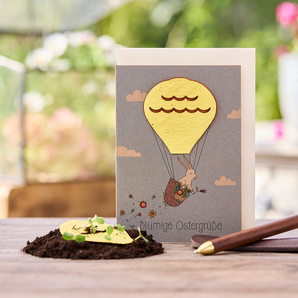 Carte de Pâques avec un lapin dans une montgolfière, à côté de lui de la terre avec des plantes en germination et un stylo.