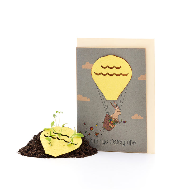 Carte de Pâques avec un lapin dans une montgolfière et du papier ensemencé en forme de cœur à partir duquel de petites plantes poussent dans le sol.
