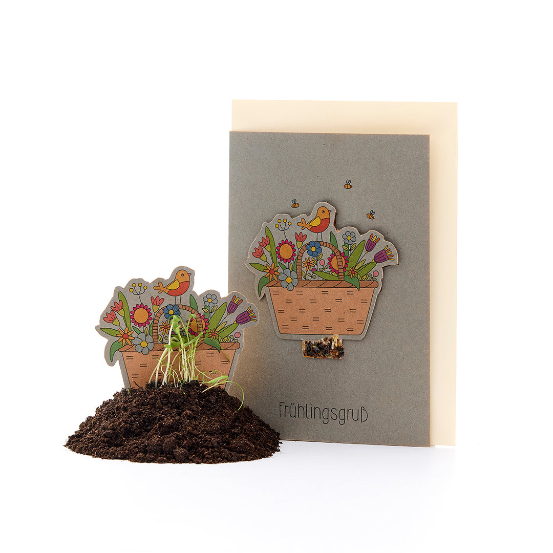 Carte de vœux avec un motif floral et un oiseau, ainsi qu'un panier de fleurs en papier dans un monticule de terre avec des plantes en train de germer, texte « Salutations de printemps ».