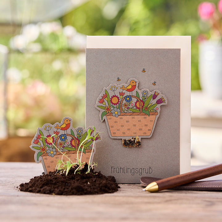 Carte de voeux avec motif printanier, panier de fleurs et oiseau, à côté un sol avec de petits semis et un stylo sur une table en bois.