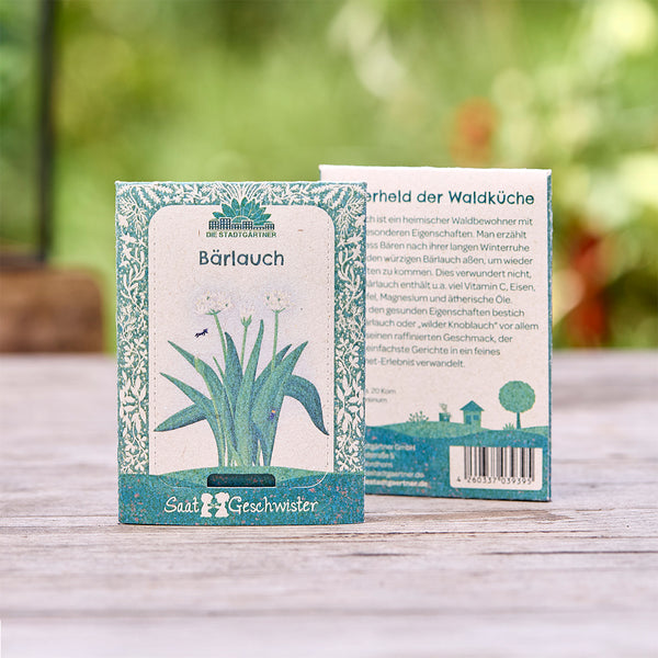 Deux sachets de graines d'ail des ours de "Die Stadtgärtner" avec illustration botanique, sur une table en bois à l'extérieur.