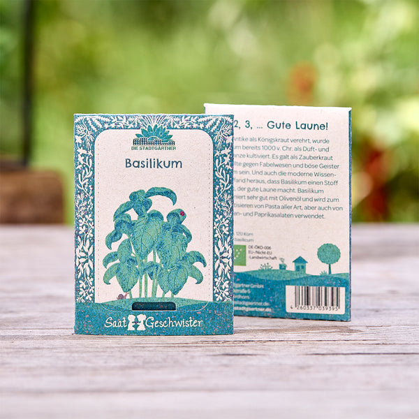 Deux sachets de graines de basilic de la marque « Die Stadtgärtner » se trouvent sur une table en bois avec un fond vert flou.