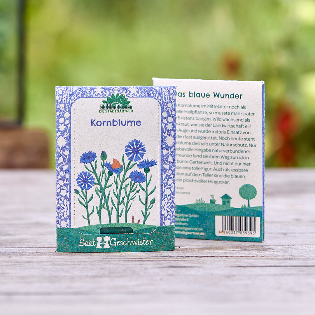 Deux sachets de graines de bleuet de la marque « Saatgeschwister » sur une table en bois, avec une illustration florale et une description du produit, et une nature floue en arrière-plan.
