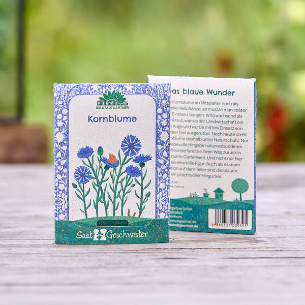 Deux sachets de graines de bleuet de la marque « Saatgeschwister » sur une table en bois, avec une illustration florale et une description du produit, et une nature floue en arrière-plan.
