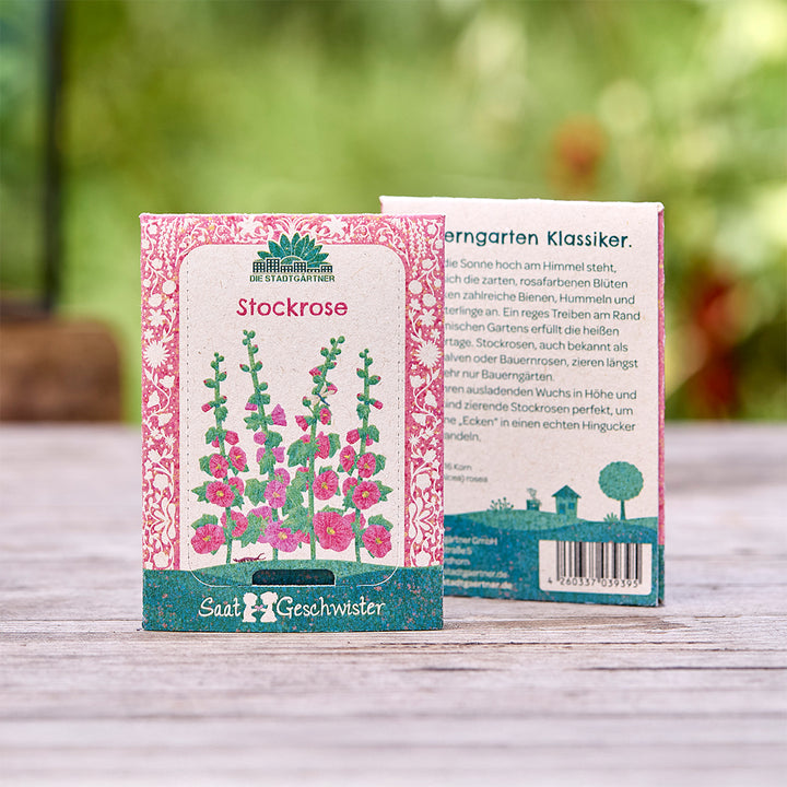 Deux sachets de graines de rose trémière avec un design coloré et un texte en allemand, placés à l'extérieur sur une table en bois.