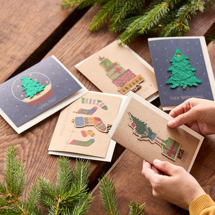 Plusieurs cartes de vœux de Noël avec des motifs colorés et des branches de sapin sur une table en bois, une carte est tenue par les mains.