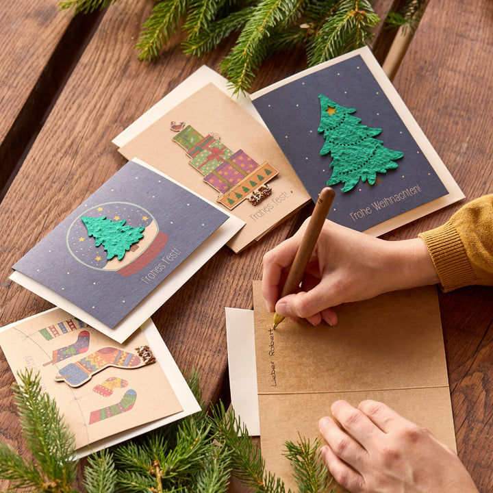 Une personne écrit une carte de Noël sur une table en bois, entourée d'autres cartes de vœux festives et de branches de sapin.
